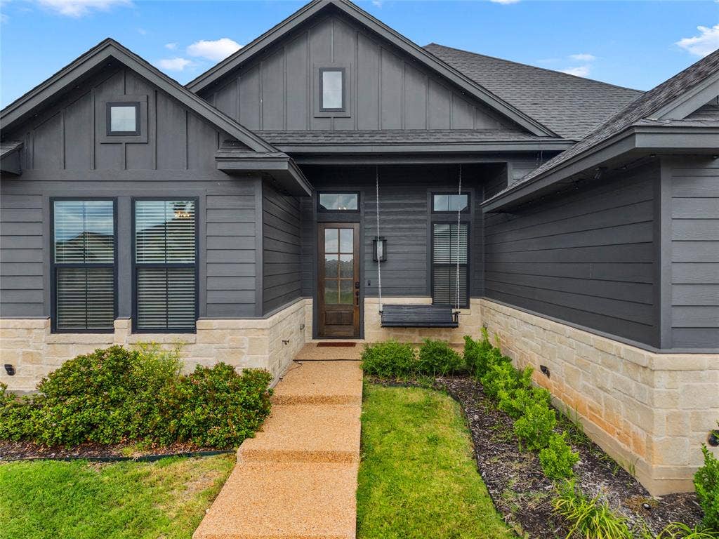 10201 Castleberry Lane, Waco, TX 76712 Architectural facade featuring dark horizontal siding and board-and-batten accents, stone veneer foundation, a gabled roofline, and an exposed aggregate walkway