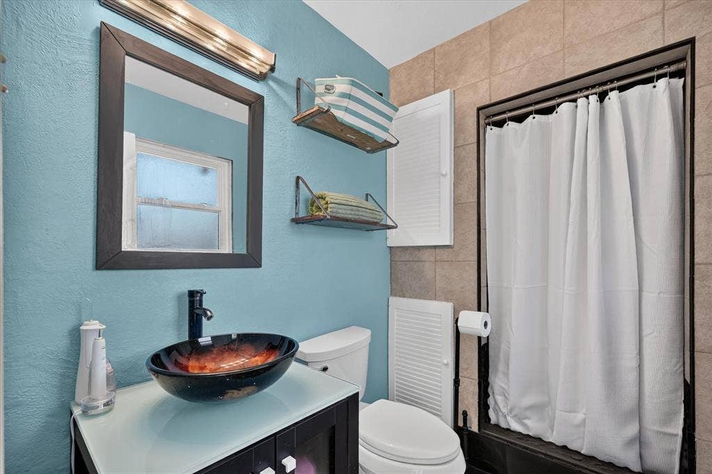 Vessel sink with a dark faucet, framed mirror, wall-mounted light fixture, and tiled shower surround