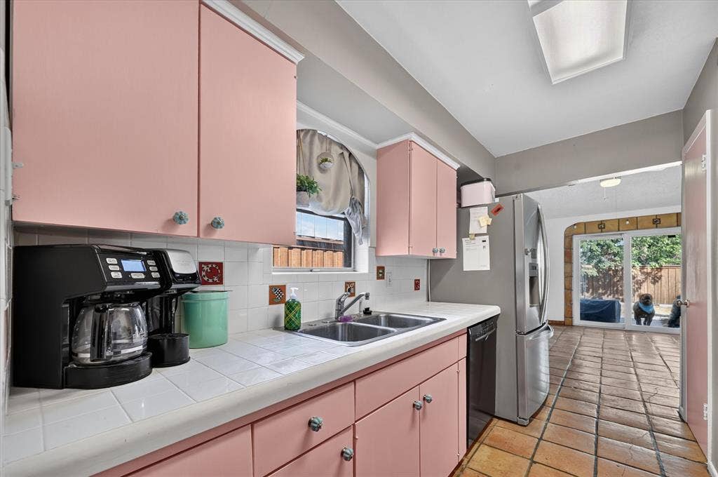 Pink cabinetry with decorative pulls, integrated dual basin sink, white tile countertops and backsplash, stainless steel refrigerator, and terracotta tile flooring