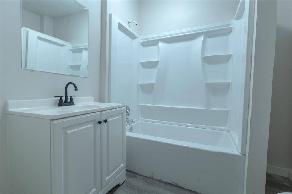 1702 Ethel Avenue, Waco, TX 76707 Modern bathroom featuring a white vanity with ample storage, ceramic sink with matte black faucet, large mirror with integrated lighting, and a bathtub-shower combination with built-in shelving and tile surround