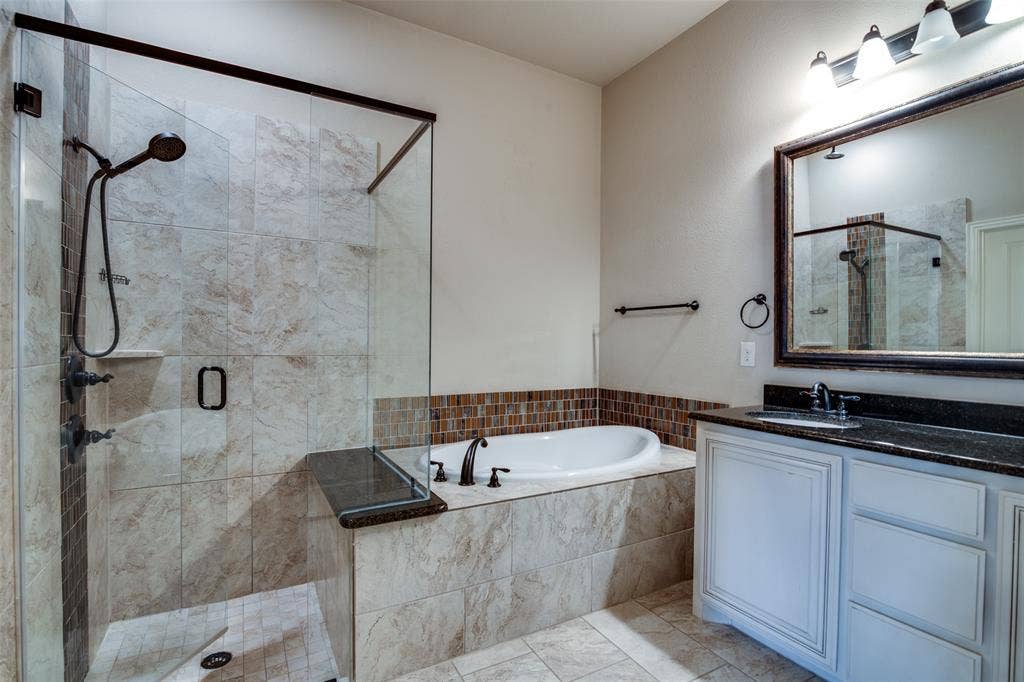 1813 Chadwick Lane, Forney, TX 75126 Glass-enclosed shower with tile surround and integrated shelving, soaking tub with tile surround and mosaic accent, single vanity with dark countertop and white cabinetry, wall-mounted mirror with decorative frame, and tile flooring