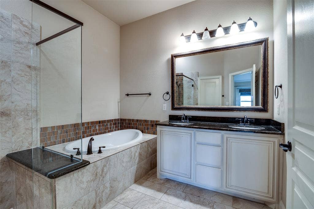 1813 Chadwick Lane, Forney, TX 75126 Dual sink vanity with dark countertop, large framed mirror, built-in soaking tub, glass-enclosed shower, and tiled flooring