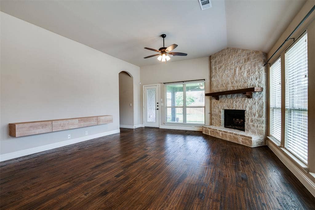 1813 Chadwick Lane, Forney, TX 75126 Spacious living area showcasing rich wood-finish flooring, ceiling fan with integrated lighting, and stone fireplace with wooden mantel