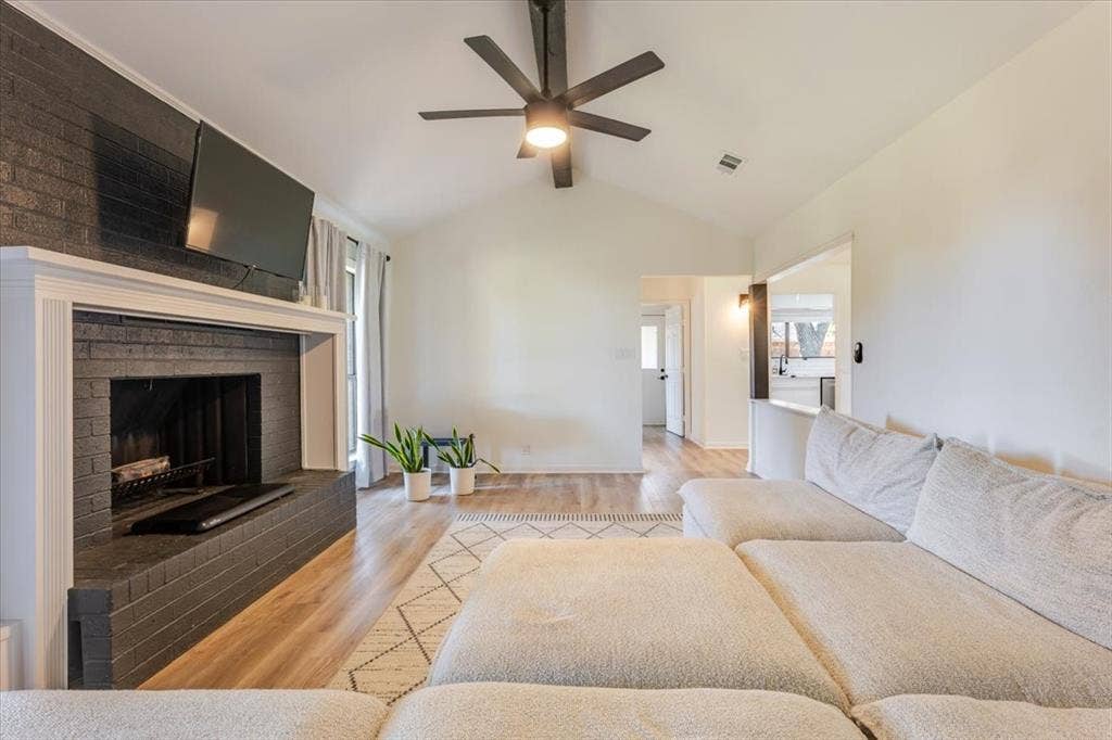 Vaulted ceiling with an installed ceiling fan, brick fireplace with white mantel, wood-finish flooring, and white walls