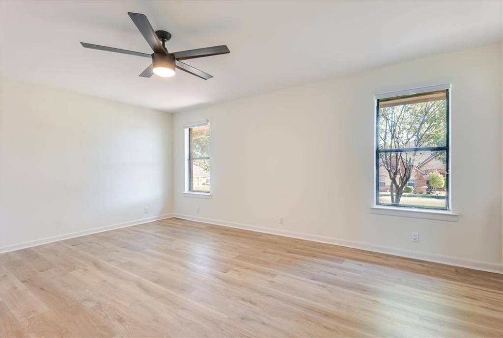 Light wood-finish flooring, two windows with white trim, and a ceiling fan with integrated lighting