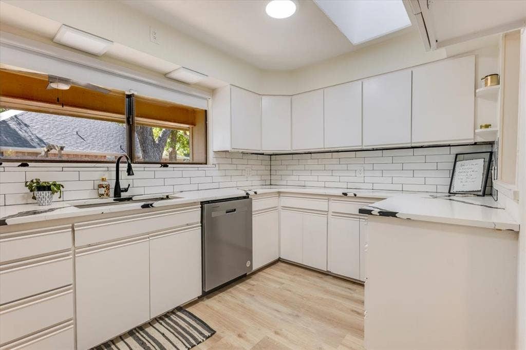 White cabinetry with black hardware, subway tile backsplash, stainless steel dishwasher, wood-finish flooring, and a skylight