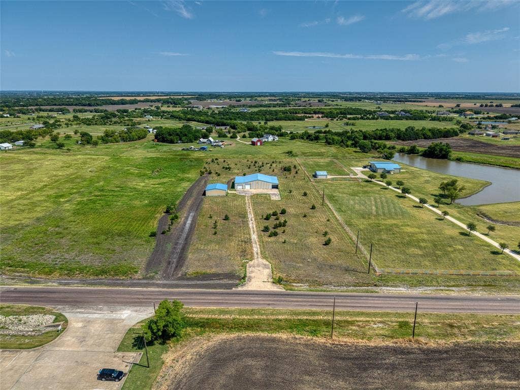 2944 CR-596, Nevada, TX 75173 Expansive property featuring multiple outbuildings with light blue metal roofs, a private pond, and a long drive leading to the main structures