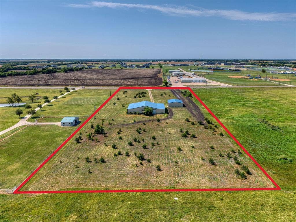 2944 CR-596, Nevada, TX 75173 Expansive land parcel featuring a primary structure with a blue metal roof, an additional outbuilding with a blue metal roof, an unpaved driveway, and scattered young trees