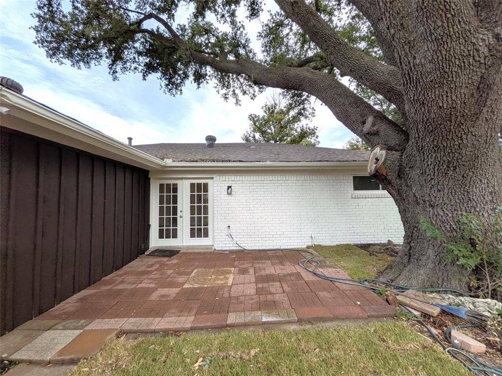 1400 Oakhill Drive, Plano, TX 75075 Brick paver patio with a French door entry, white painted brick facade, brown vertical siding, and an established mature tree