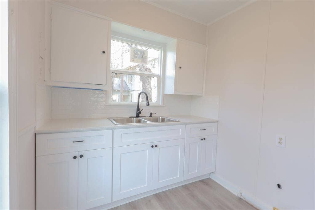 1704 Ethel Avenue, #B, Waco, TX 76707 White shaker-style cabinetry with dark hardware, dual stainless steel sinks with a gooseneck faucet, light-toned countertops, white subway tile backsplash, and wood-finish flooring
