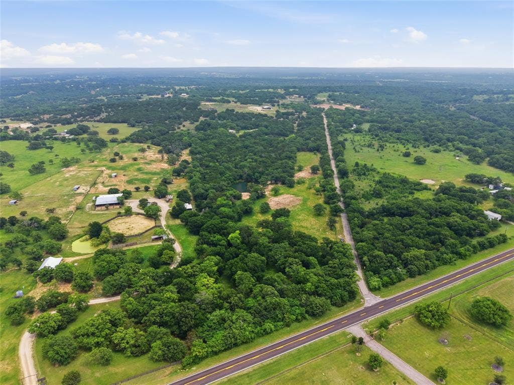 TBD Manley Road, Weatherford, TX 76088 Expansive aerial landscape featuring a paved road, dense tree coverage, open green fields, a visible pond, and multiple structures