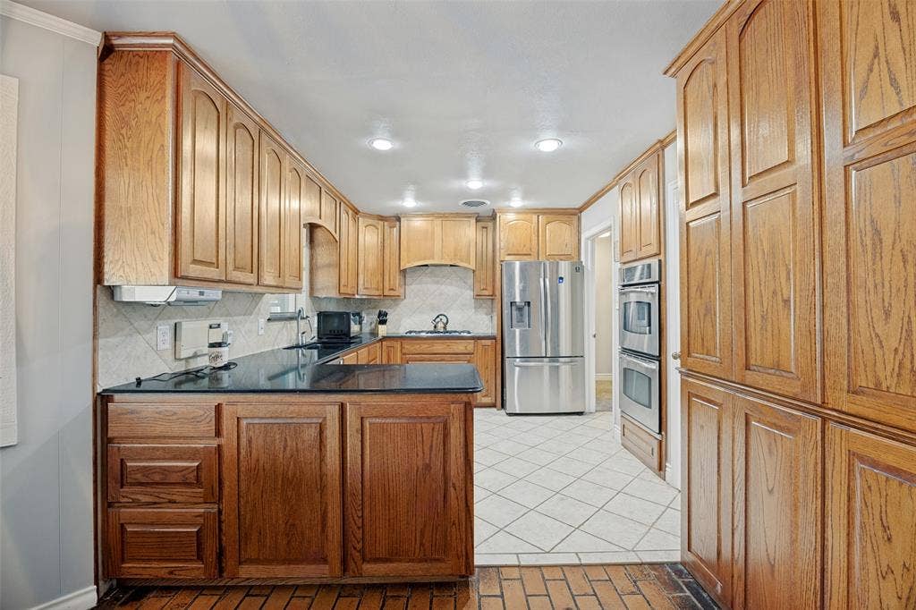 6401 Stonewall Street, Greenville, TX 75402 Kitchen featuring wood-finish cabinetry, dark countertops, a tiled backsplash, and stainless steel appliances including a refrigerator and double ovens