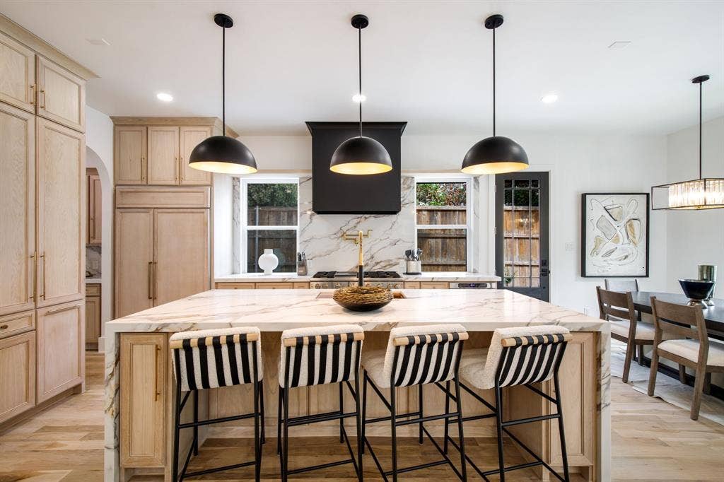 Spacious kitchen featuring light wood-finish cabinetry, a large kitchen island with a stone countertop, three pendant lights, and a built-in range hood