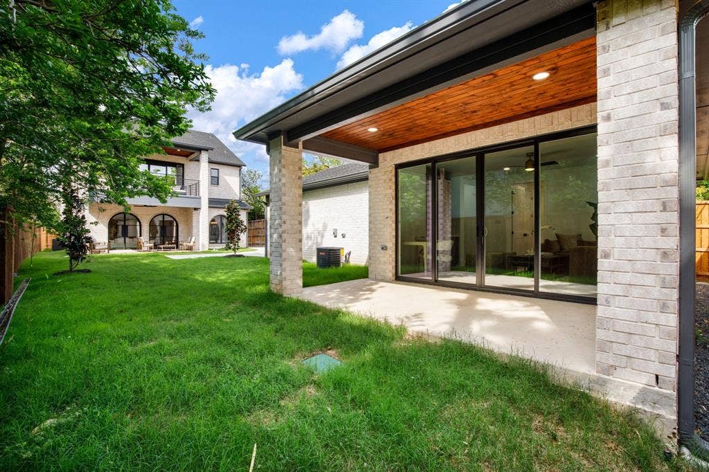 Modern residence featuring an expansive manicured lawn with mature landscaping, extensive glass sliding doors opening to the rear patio, white brick exterior, wood-finished soffit detail, and in-ground hardscaping throughout the backyard entertaining space