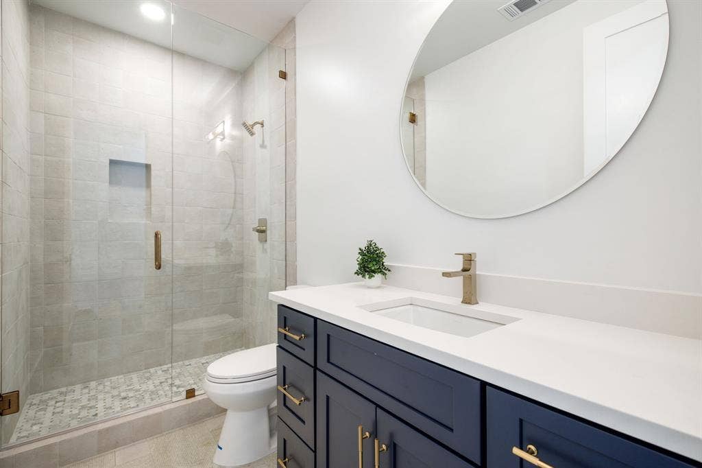 Glass-enclosed shower with tiled walls and floor, vanity with white countertop and integrated sink, dark blue cabinetry with brass hardware, and a large oval mirror