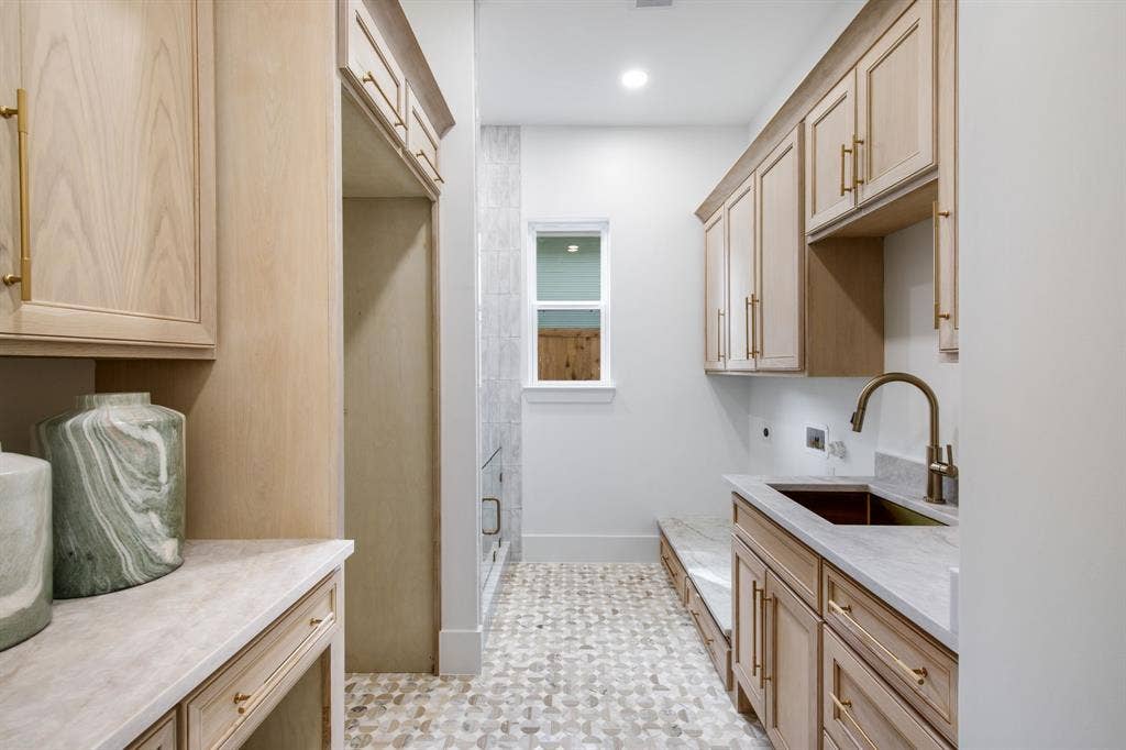 Light wood-finish cabinetry with gold-tone hardware, undermount sink with a gooseneck faucet, light-toned countertop, and mosaic tile flooring