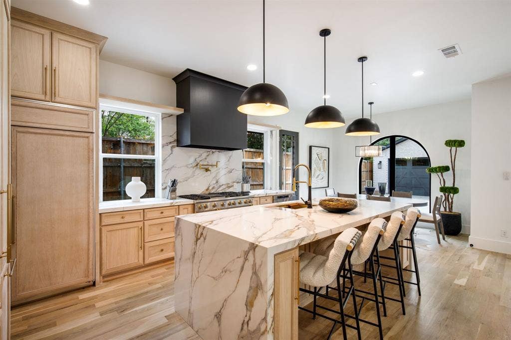 Gourmet kitchen with a waterfall island, integrated appliances, natural wood-finish cabinetry, and a black range hood