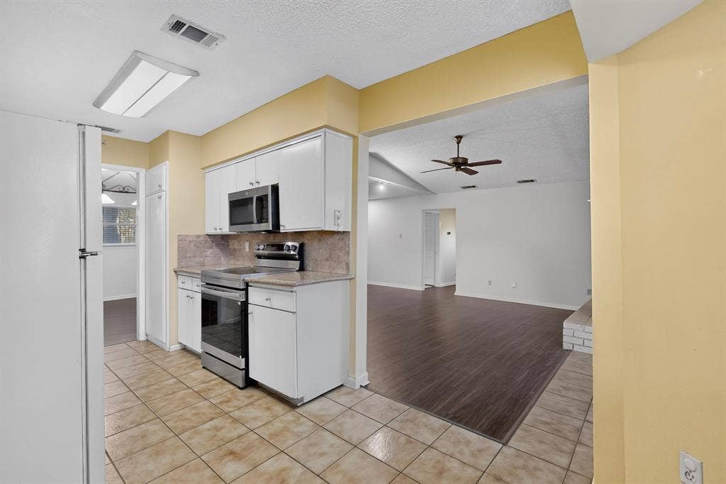 Kitchen featuring white cabinetry, stainless steel appliances, tile flooring, and a pass-through opening to a living area with wood-finish flooring and a ceiling fan