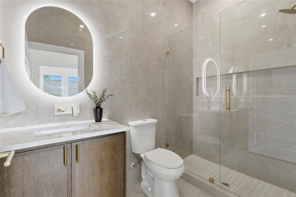 3424 Navaro Street, Dallas, TX 75212 Illuminated oval mirror, wood-finish vanity with gold-tone hardware, white countertop with integrated sink, glass-enclosed shower, and full-height wall tiling