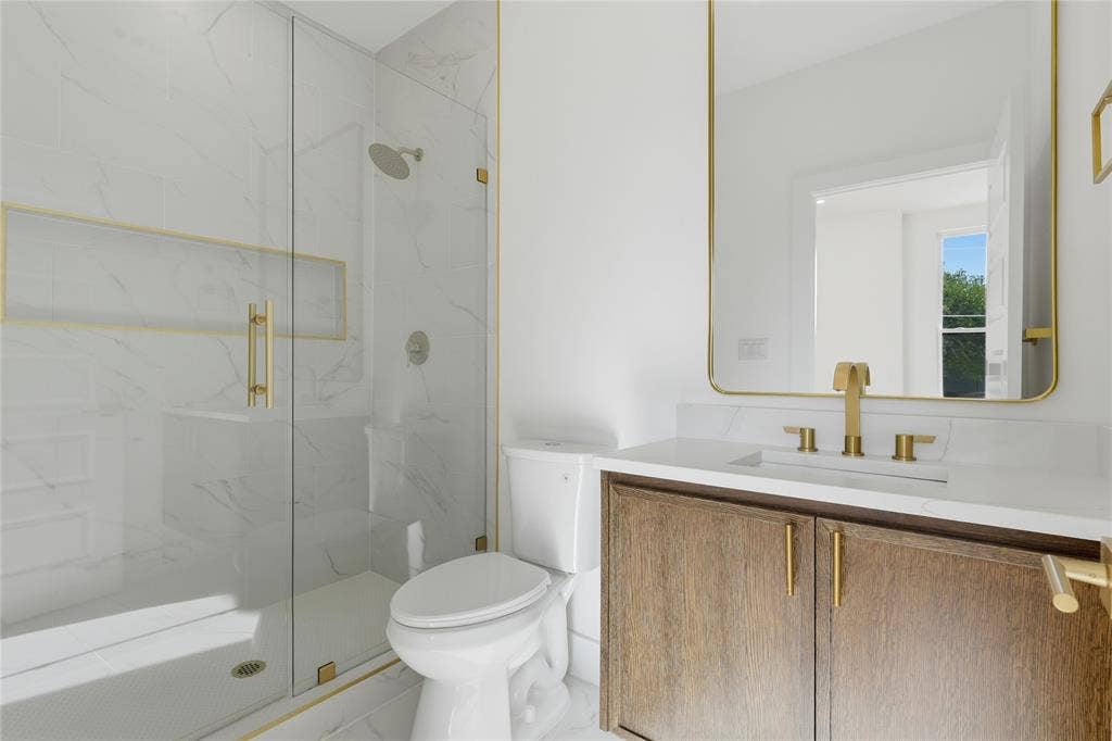 3424 Navaro Street, Dallas, TX 75212 Walk-in shower with glass enclosure and gold-finish hardware, white tile with subtle veining, wood-finish vanity with gold-finish pulls, gold-finish faucet, and a rectangular mirror with a gold-finish frame