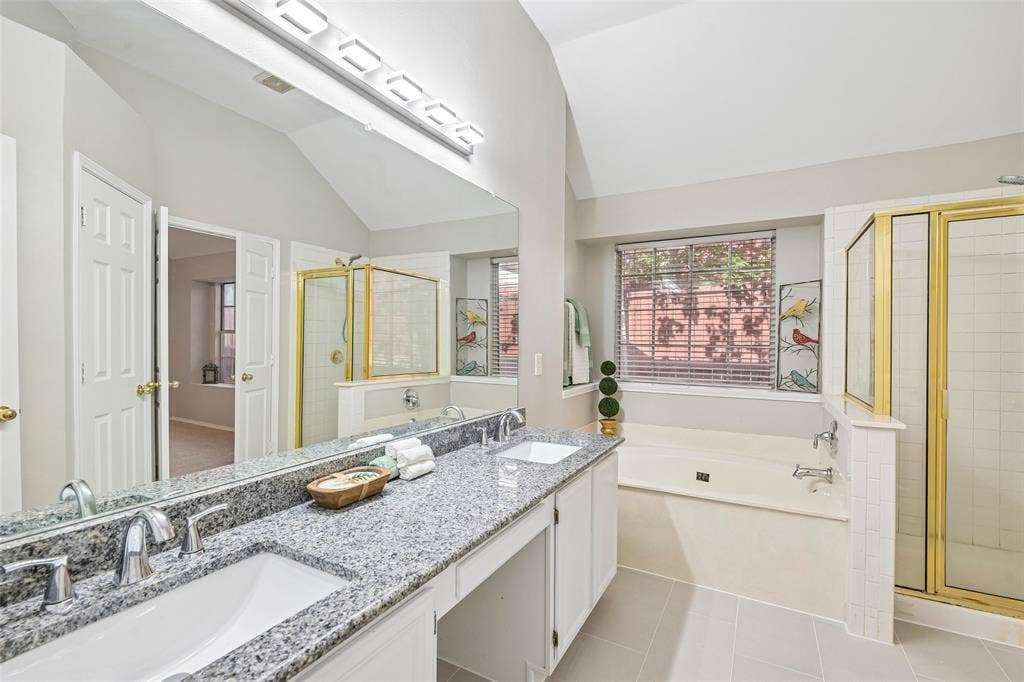 Dual vanity with granite countertops, an undermount tub, a separate shower with a gold-framed enclosure, and light-toned floor tiles