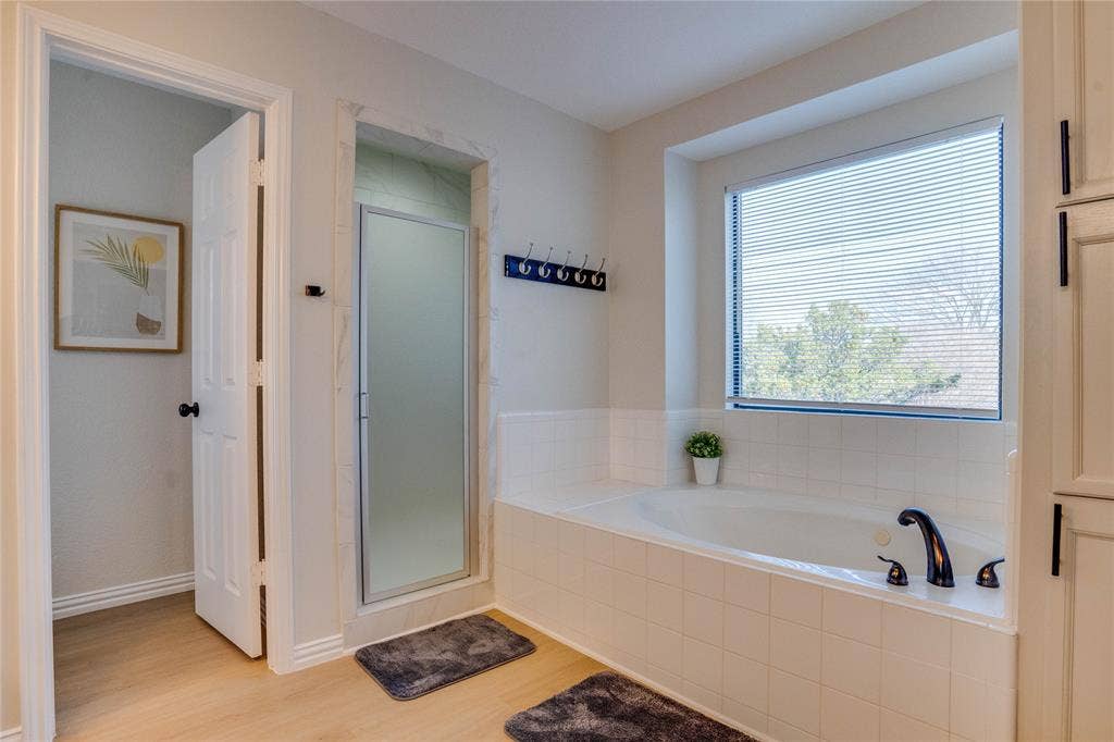 6003 Lloyd Court, Dallas, TX 75252 Spa-inspired primary bathroom featuring a soaking tub with deck-mounted faucet, wood-finish flooring, and abundant natural light from a large picture window