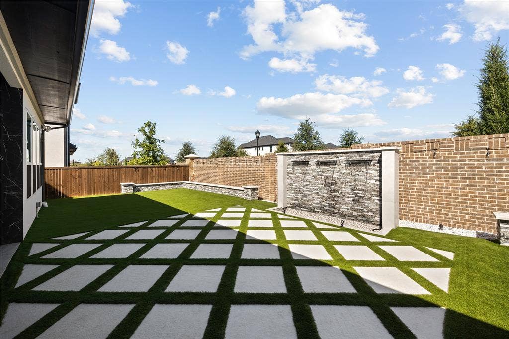 12512 Deer Trail, Frisco, TX 75035 Expansive backyard featuring a water feature with stone-finish cladding, a brick perimeter wall, and a geometric pattern of concrete pavers set in artificial turf