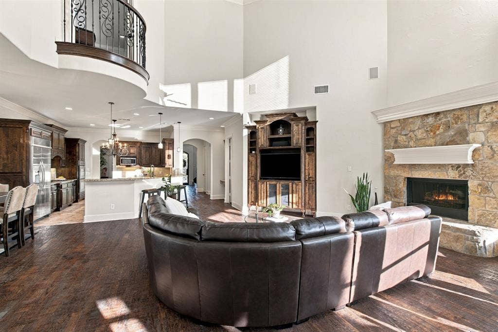 Two-story living area featuring wood-finish flooring, a full-height stone fireplace with a decorative mantel, custom built-in cabinetry, and a curved balcony with iron railing
