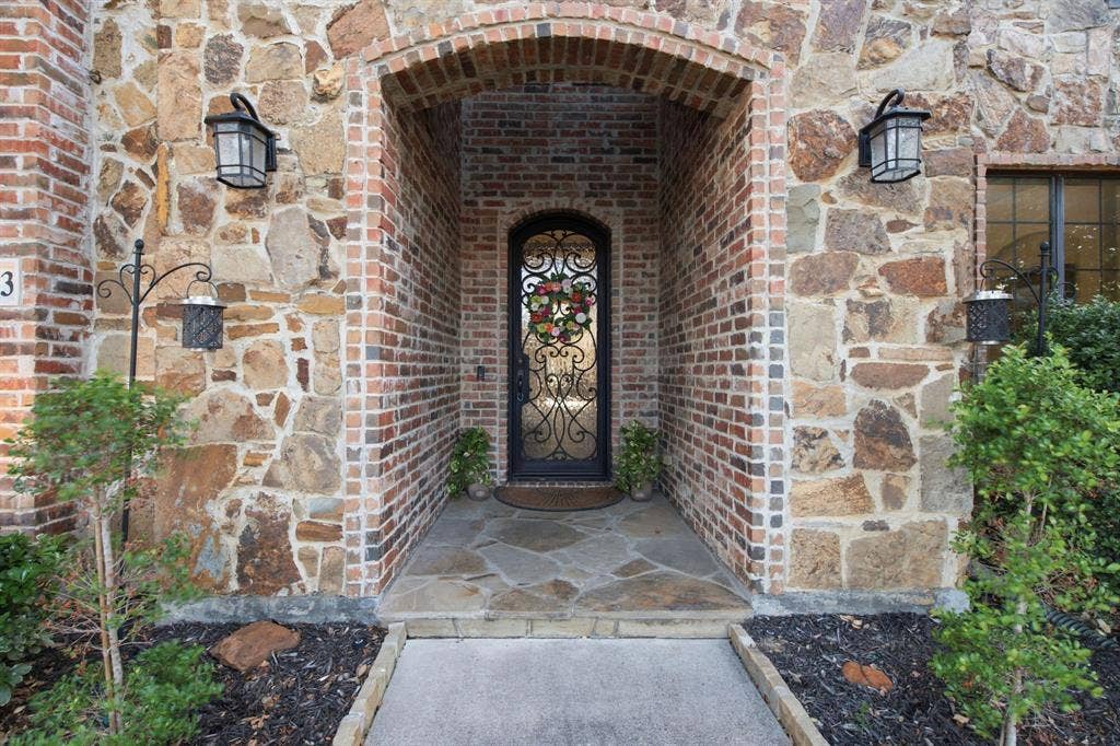 Exterior entrance featuring custom iron and glass arched door, multi-tonal stone facade, brick-accented arched entryway, and flagstone porch flooring