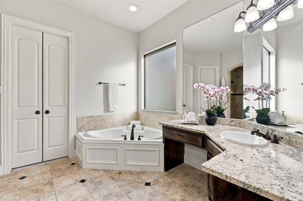 Corner soaking tub with tile surround, stone-finish vanity countertop with an integrated oval basin, full-width vanity mirror, dark wood-finish cabinetry, and ceramic tile flooring with decorative inlay accents