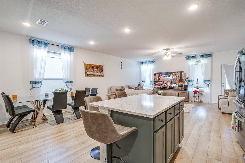 Open-concept living space featuring a large kitchen island with storage cabinetry and seating, wood-finish flooring throughout, recessed ceiling lighting, and multiple windows with blue-toned window treatments providing natural light