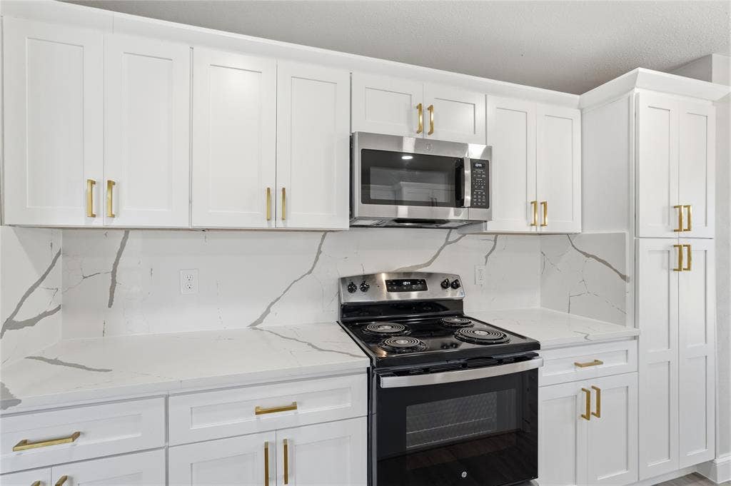 5514 Round Rock Road, Garland, TX 75044 White shaker-style cabinetry with gold-tone hardware, white countertops, coordinating white backsplash with gray veining, stainless steel microwave, and built-in electric range