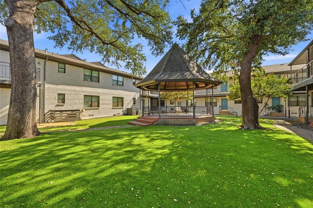 Lush green lawn with mature trees, a central gazebo with a shingled roof, and brick-clad buildings