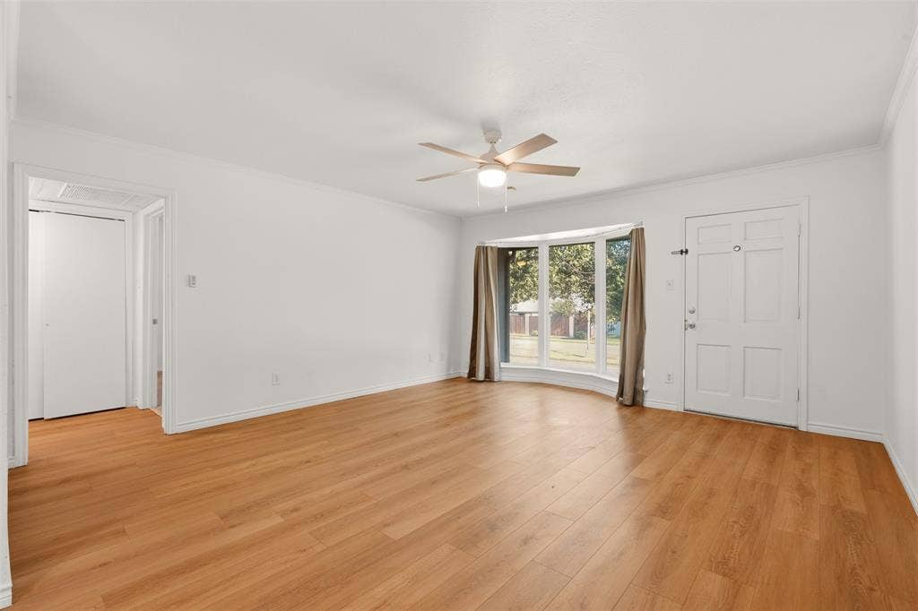 Wood-finish flooring throughout, bay window with multiple panes, ceiling fan light fixture, white interior door, and a white front entry door