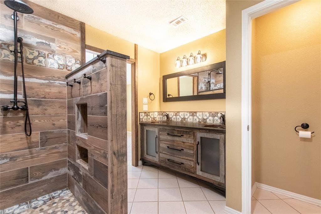 Tiled shower with a rain shower head, dual vanity with undermount sinks, patterned backsplash, and wood-finish cabinetry