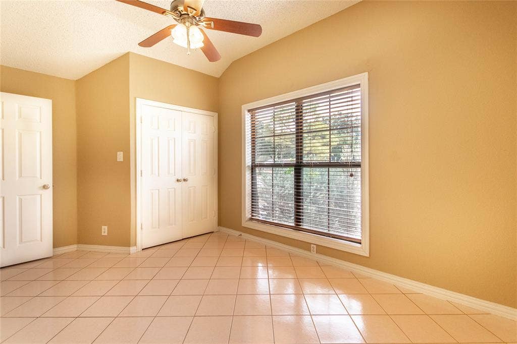 Tile flooring, vaulted ceiling, ceiling fan with light fixture, window with blinds, and bi-fold closet doors
