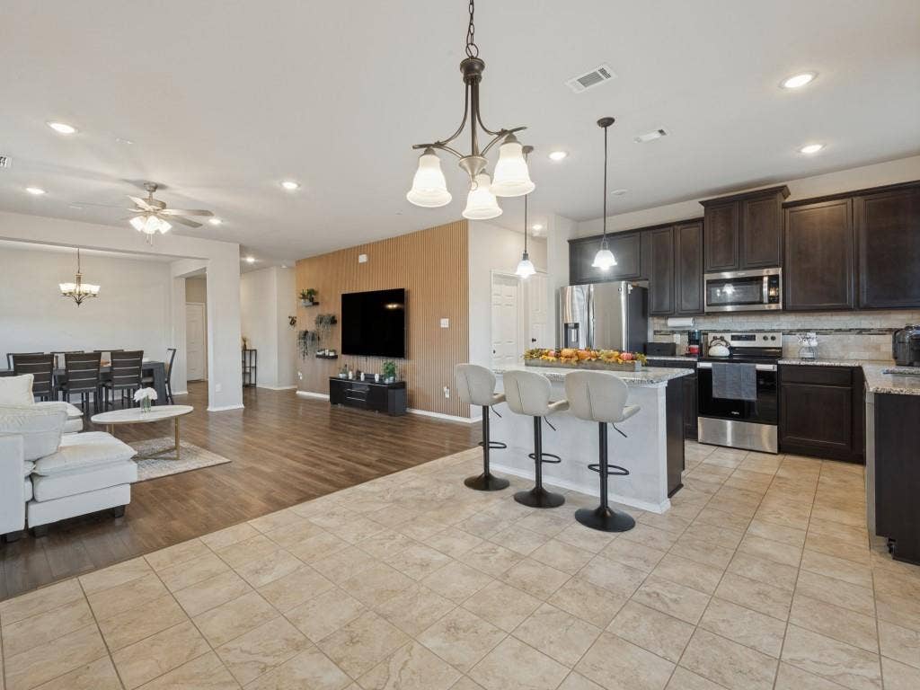 3825 S Courtney Court, Heartland, TX 75126 Open-concept living area featuring wood-finish flooring throughout, a centered kitchen island with seating for multiple guests, stainless steel appliances, dark cabinetry with contrasting countertops, pendant lighting, recessed ceiling lights, and a ceiling fan