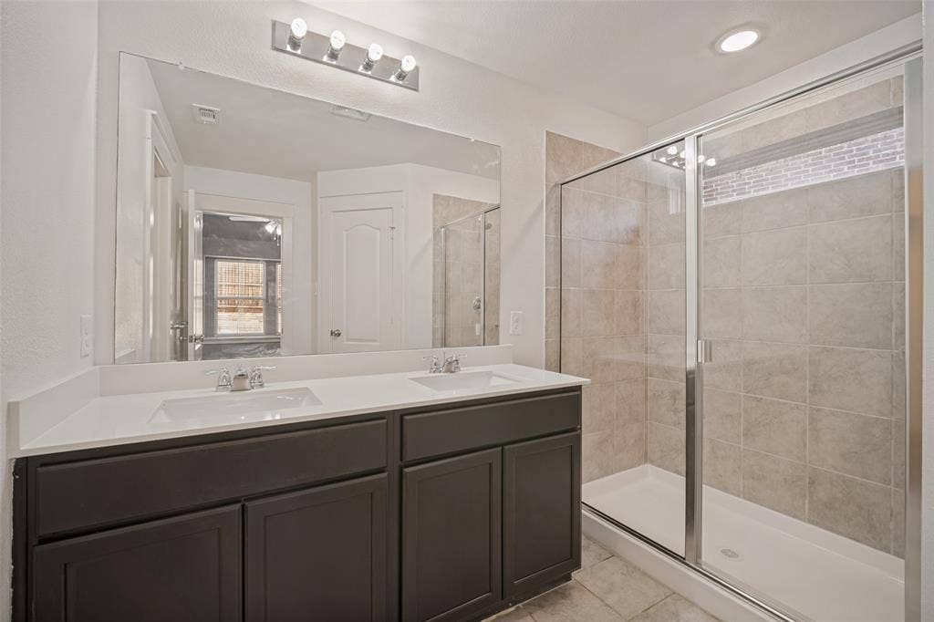 2324 Neff Lane, Forney, TX 75126 Dual-sink vanity with a solid-surface countertop and charcoal-finish cabinetry, featuring a frameless-style mirror, chrome hardware, and a walk-in shower with tiled surround