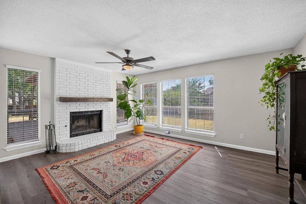 519 Fox Creek Court, Denton, TX 76209 White-painted brick fireplace with a wood mantle, ceiling fan, dark wood-finish flooring, and multiple windows