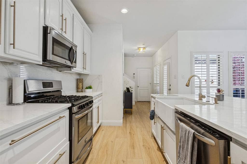 6816 Livorno Lane, Frisco, TX 75034 Custom white shaker cabinetry with gold-tone hardware, stainless steel appliances, white countertop surfaces, wood-finish flooring, and a farmhouse-style sink with a gold-tone faucet