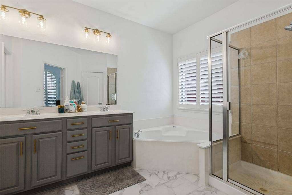 6816 Livorno Lane, Frisco, TX 75034 Primary ensuite bathroom has dual vanity with gray cabinetry and gold-tone hardware, large wall mirror, recessed corner bathtub, and walk-in shower with tile surround. newly tiled bathroom floor