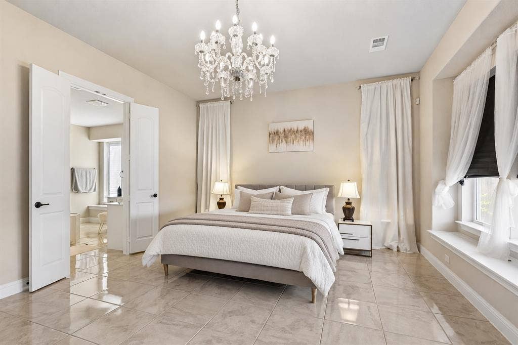 3613 Sparkling Drive, Plano, TX 75074 Elegant primary bedroom suite featuring soaring ceilings with crystal chandelier, tile flooring throughout, dual ensuite access via French doors, floor-to-ceiling windows with custom drapery, paired nightstands with integrated lighting, and a spacious layout accommodating a substantial bed frame