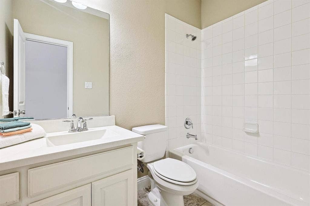 White vanity with integrated sink and chrome faucet, large frameless mirror, white bathtub with a tiled surround, and a white toilet