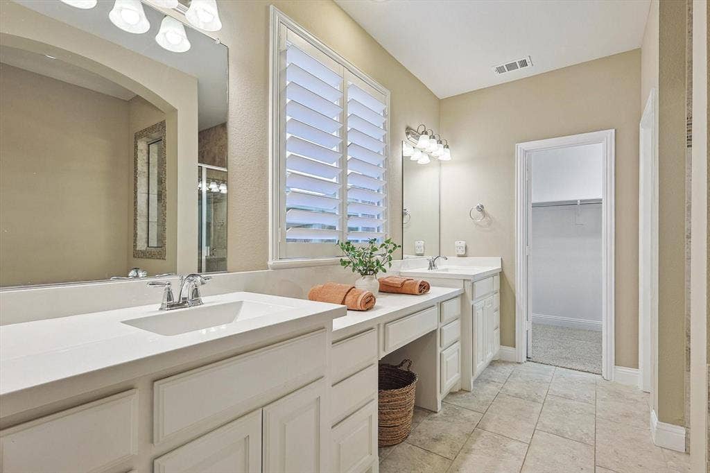 Dual vanity with white cabinetry and integrated sinks, a built-in makeup area, large window with plantation shutters, and tiled flooring