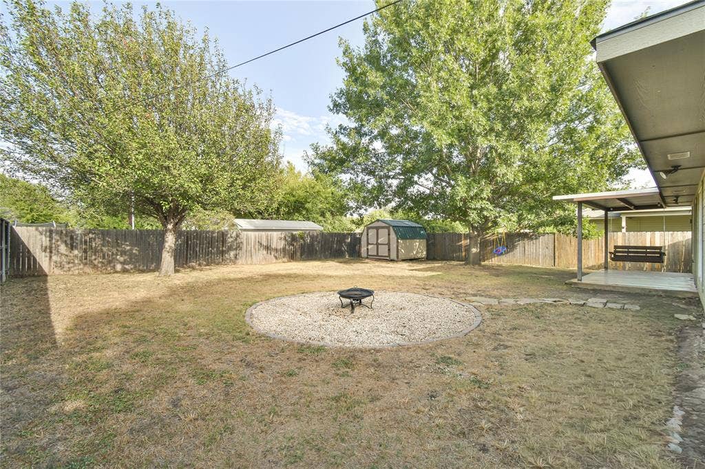 2108 8th Street, Brownwood, TX 76801 Expansive backyard featuring a privacy fence, mature trees, a gravel fire pit area, a covered patio, and a storage shed