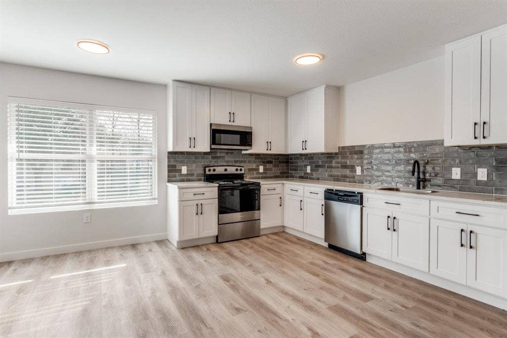 5917 Gaston Avenue, #115, Dallas, TX 75214 White cabinetry with black hardware, stainless steel appliances, dark subway tile backsplash, light wood-finish flooring, and a large window with blinds