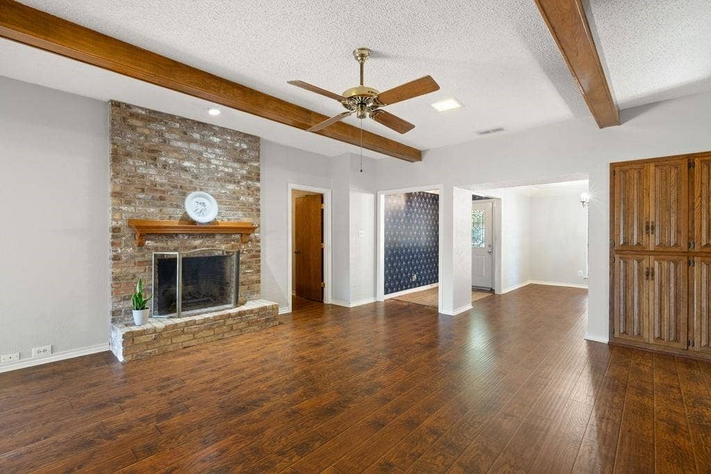 Brick-faced fireplace with wood mantel, wood-finish flooring, exposed ceiling beams, and a ceiling fan with integrated lighting