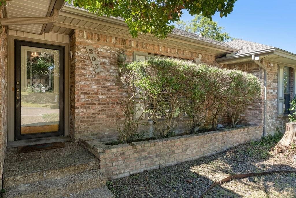 Brick exterior with a storm door, concrete steps, and a brick planter box with established shrubs