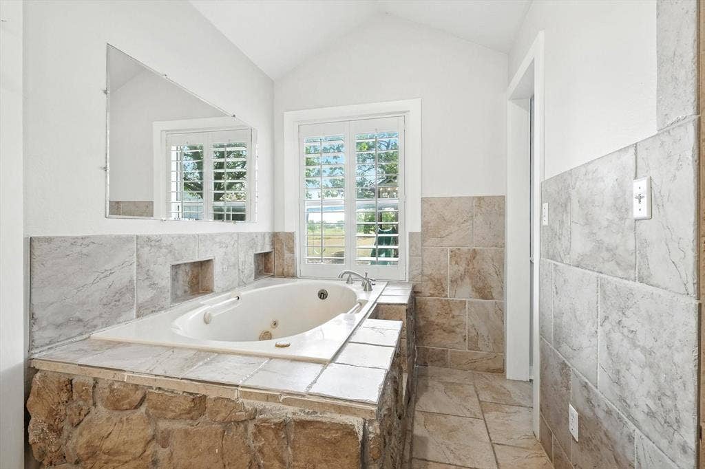 185 Quail Run Court, Decatur, TX 76234 Built-in jetted tub surrounded by stone and tile, featuring a mounted mirror, vaulted ceiling, and plantation-shuttered windows