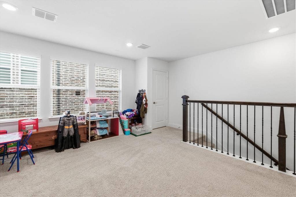 2637 Basswood Drive, Northlake, TX 76226 Carpeted room featuring three windows with blinds, a white door, and a staircase with dark wood railings and black iron balusters